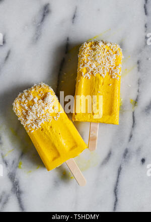 Blick von oben auf die hausgemachte zwei Eis am Stiel aus Kokosmilch und Gefrorenes Mango über weißen Marmor. Gesunde veganes Essen und Sommer Konzept. Stockfoto