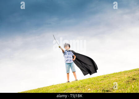 Stolz, Jungen spielen der super Held tragen einen selbstgemachten Umhang und mit einem Spielzeugschwert auf die Skyline vor blauem Himmel Stockfoto
