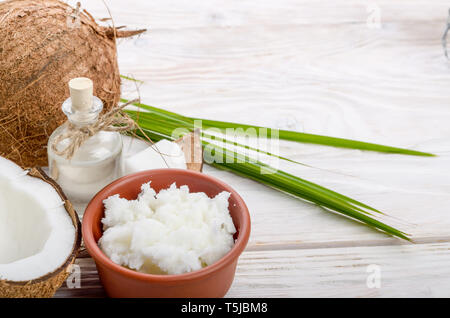 Hintergrund der Kokosnuss, Kokosnussschalen, Öl in Ton Schüssel und Glas auf weissem Holztisch Stockfoto