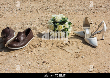 Braut und Bräutigam Schuhe und handabdrücke am Strand im Sand mit Hochzeit Ringe und Blumenstrauß Stockfoto