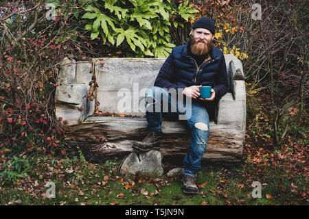 Bärtiger Mann mit schwarzen Mütze sitzend auf Bank aus Holz im Garten, Holding blau becher, an der Kamera schaut. Stockfoto
