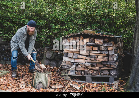 Der bärtige Mann mit schwarzer Mütze und Parka im Garten im Herbst, mit ax Stück Holz auf hackklotz zu hacken. Stockfoto