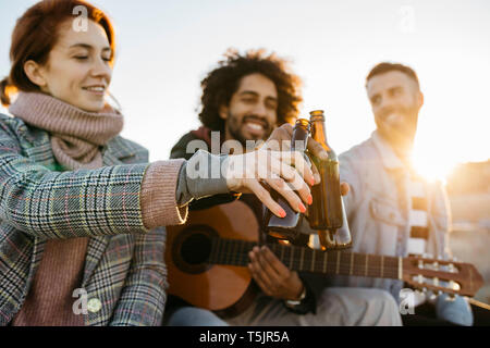 Drei glückliche Freunde mit Gitarre toasten Bierflaschen bei Sonnenuntergang Stockfoto