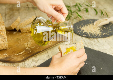 Nahaufnahme von Frau Hände gießen natives Olivenöl extra aus einer Ampulle auf einer Scheibe Brot im rustikalen Schuß Zusammensetzung Stockfoto
