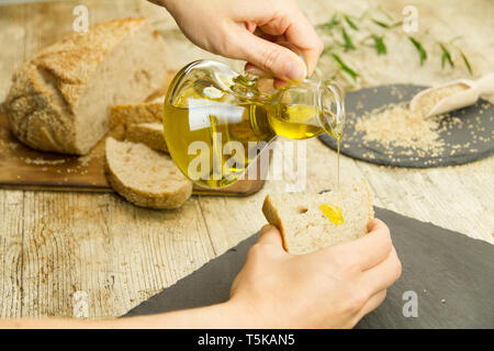 Nahaufnahme von Frau Hände gießen natives Olivenöl extra aus einer Ampulle auf einer Scheibe Brot im rustikalen Schuß Zusammensetzung Stockfoto