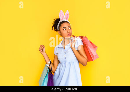 Junge Frau mit cute dress und hasenohren kopfband stehend auf gelben Hintergrund holding Einkaufstaschen beiseite, isoliert, neugierig Stockfoto
