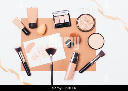 Schönheit Produkte und Zubehör für ebenmäßigen Teint, Ansicht von oben. Dekorative Kosmetik und Make-up-Pinsel mit beige Seidenband auf weißem Hintergrund. Stockfoto