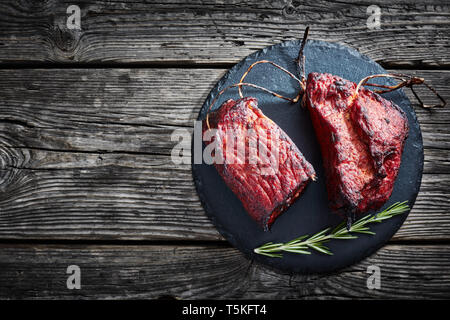 Zwei Stücke von geräuchertem Grill Schweinefilet mit Garn und frischem Rosmarin auf einem runden Stein Platte auf einem rustikalen Holztisch, horizontale Ansicht von ab Stockfoto
