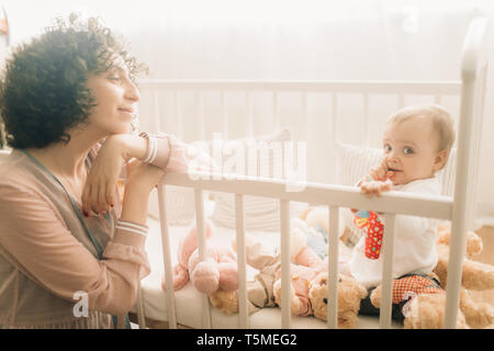 Glückliche Mutter sitzt neben Ihr Baby mit Spielzeug in die Krippe. Stockfoto