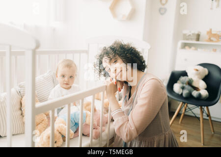 Glückliche Mutter sitzt neben Ihr Baby mit Spielzeug in die Krippe. Stockfoto