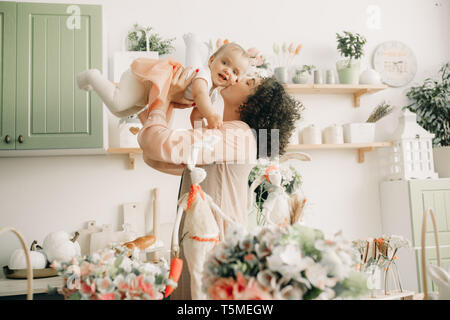 Glückliche Mutter spielt und küsst ihr Baby in der Küche auf dem Hintergrund von Ostern Dekorationen. Stockfoto