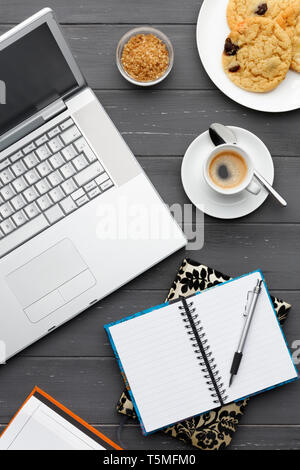 Hochformat eine Kaffeepause mit Laptop, Kaffee, Kekse, Zucker und mehrere Notizblöcke auf einem dunklen Hintergrund Stockfoto