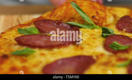 Appetitlich frisch Hot peperoni Pizza auf einen hölzernen Tisch dekorieren mit grünen durch eine unscharfe Abbildung der Mann oder die Frau Koch auf Hintergrund in Zeitlupe Stockfoto