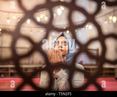 Blick durch Zaun: muslimische Frau in Kopftuch und Hijab betet mit den Händen in der Luft in Moschee. Religion beten Konzept. Stockfoto