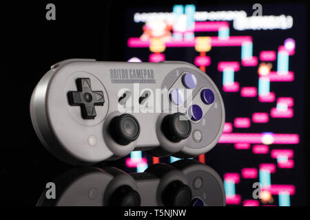 Ein grauer, drahtlose Spiele Controller, aufrecht sitzend auf einer gespiegelten Oberfläche, vor einem Out-of-focus arcade spiel. Stockfoto