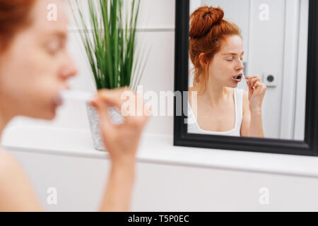 Junge rothaarige Frau, die ihre Zähne putzen in einem weißen Badezimmer in einem über die Schulter Blick auf ihr Spiegelbild im Spiegel