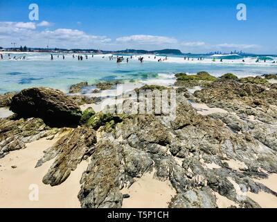 Suchen von den Felsen sonnigen Strand mit kristallklarem türkisfarbenem Wasser. Surfer und Schwimmer im Hintergrund Schwimmen in den schönen Gewässern. Stockfoto