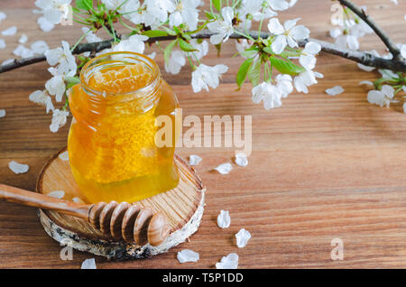 Honig Glas auf Scheibe Birkenholz mit Wabe und Honig Pendelarm auf Holzuntergrund mit Kirschblüten. Stockfoto