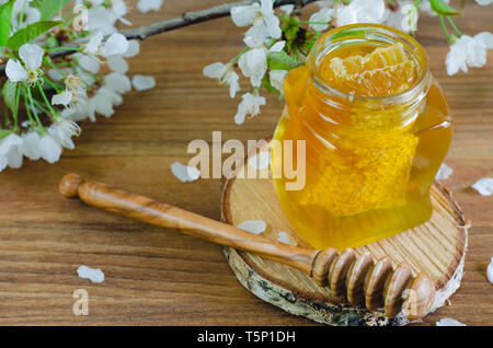 Honig Glas auf Scheibe Birkenholz mit Wabe und Honig Pendelarm auf Holzuntergrund mit Kirschblüten. Stockfoto
