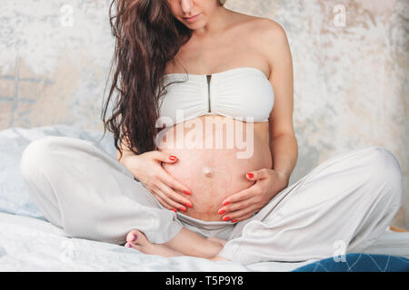 Foto zuschneiden der schönen schwangeren Mädchen Junge Frau mit langen, lockigen Haar sitzt im Lotussitz auf dem Bett. Schwangerschaft Yoga und Natur ethnischen Schönheit Konzept Stockfoto