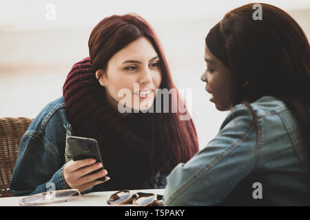 Zwei junge schöne Mädchen sitzen an einem Tisch und reden über das Handy. Stockfoto