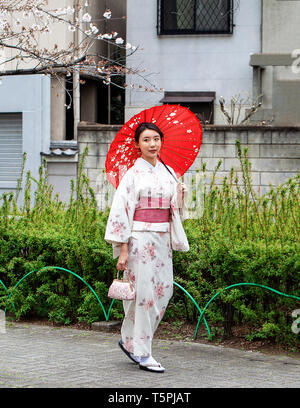 Schönen japanischen Frau in weißem Kimono mit roter Schärpe und roten Regenschirm in einem Park neben einem Kirschblüte Baum Stockfoto