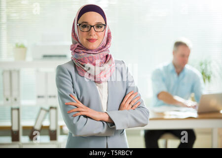 Schöne junge berufstätige Frau im Hijab, Anzug und Brille in Büro, lächelnd. Portrait von zuversichtlich muslimischen Geschäftsfrau. Moderne Büro wit Stockfoto