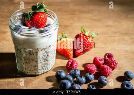 Jam jar über Nacht Hafer und Joghurt im Glas Marmeladenglas auf rustikalen Holzbank mit frischen Erdbeeren, Himbeeren und Heidelbeeren Stockfoto