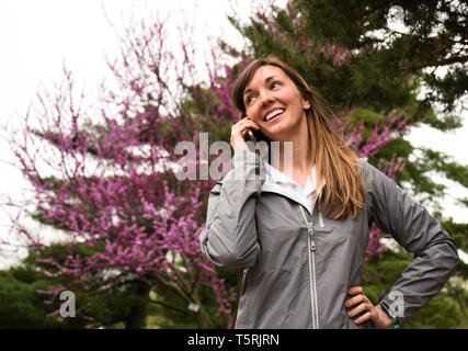 Attraktive, lächelnden jungen Frau, die sich in der Zelle Handy, die Hände auf den Hüften, im Stadtpark, rosa Baum Hintergrund, grünes Gras, bewölkten Tag - Bild Stockfoto