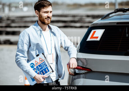 Porträt einer stattlichen Treiber Ausbilder stehend mit Beschilderung in der Nähe des Lernens Auto auf dem Trainingsgelände im Freien Stockfoto