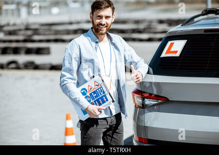 Porträt einer stattlichen Treiber Ausbilder stehend mit Beschilderung in der Nähe des Lernens Auto auf dem Trainingsgelände im Freien Stockfoto