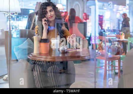 Schöne Frau am Telefon zu sprechen, während an einem Tisch in einem Cafe hinter dem Glas sitzen Stockfoto
