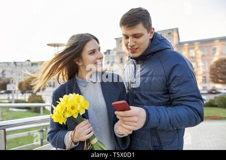 Outdoor Portrait von schöne romantische Paare, junge Mann und Frau mit Blumenstrauß aus gelben Blüten der Narzissen und Suchen in Smartphone, Spring City Stockfoto