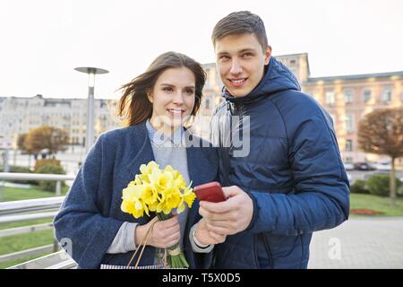 Outdoor Portrait von schöne romantische Paare, junge Mann und Frau mit Blumenstrauß aus gelben Blüten der Narzissen und Suchen in Smartphone, Spring City Stockfoto