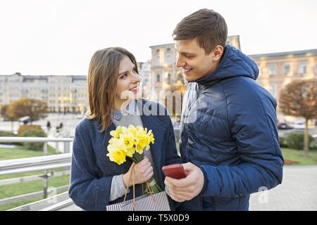 Outdoor Portrait von schöne romantische Paare, junge Mann und Frau mit Blumenstrauß aus gelben Blüten der Narzissen und Suchen in Smartphone, Spring City Stockfoto