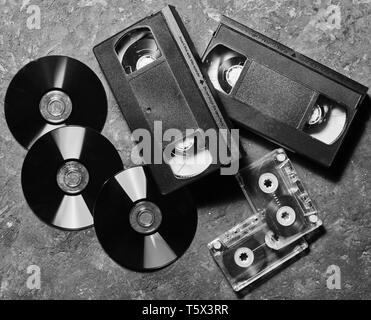 Unterhaltung und Medien Technologie aus den 90er Jahren. Cds, Audiokassetten, Videokassetten auf einem schwarzen Betonoberfläche. Ansicht von oben. Pop Licht. Stockfoto