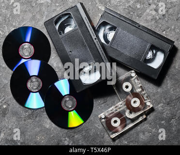 Unterhaltung und Medien Technologie aus den 90er Jahren. Cds, Audiokassetten, Videokassetten auf einem schwarzen Betonoberfläche. Ansicht von oben. Pop Licht. Stockfoto
