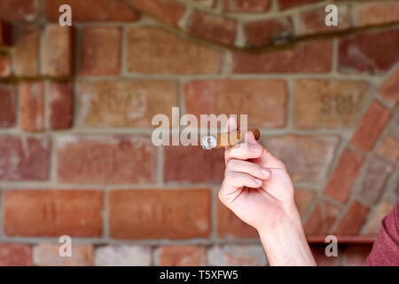 Ein junger Mann hält eine kubanische Zigarre in der Hand und raucht Bei einer unscharfen Mauer Hintergrund Stockfoto