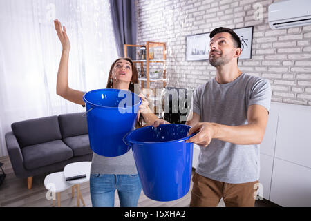Junge Mann und Frau mit blauen Eimer sammeln Wasser beschädigte Decke im Wohnzimmer Stockfoto