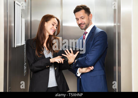 Lächelnde junge Unternehmerin und Unternehmer diskutieren während der Verwendung digitaler Tablet in der Nähe des Aufzugs, Stockfoto