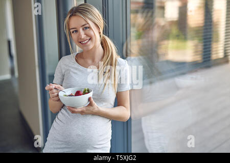 Glückliche gesunde schwangere junge blonde Frau, lehnte sich gegen eine Terrasse Fenster lächelt in die Kamera und genießen Sie eine Schale mit frischem Obstsalat Stockfoto