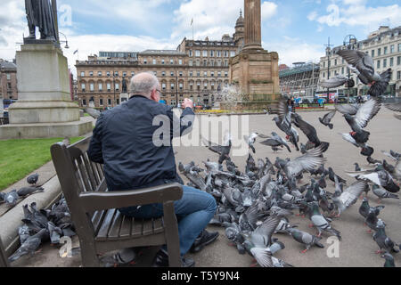 Glasgow, Schottland, Großbritannien. 27.April 2019. UK Wetter. Ein Mann sitzt auf einer Bank, die Fütterung der Tauben auf einem sonnigen Nachmittag auf dem George Square. Credit: Skully/Alamy leben Nachrichten Stockfoto