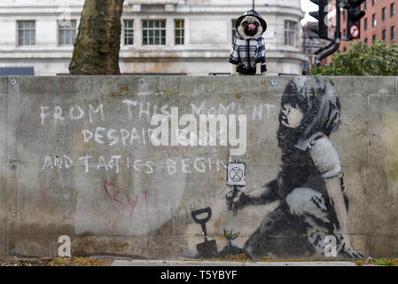 London, Großbritannien. 27. April 2019. Lilie der Mops sitzt neben einem Kunstwerk, das an einer Wand im Marble Arch nach zehn Tagen der Proteste in London vom Aussterben Rebellion, eine Gruppe fordert, dass Regierungen Maßnahmen zur Bekämpfung des Klimawandels erschienen ist. Jetzt in einer Schutzhülle aus Kunststoff abgedeckt, das Kunstwerk ist der gefeierte Street Artist Banksy zugeschrieben worden und stellt ein Bild von einer Pflanze und ein Mädchen, dass eine Gartengeräte mit dem Aussterben Rebellion Logo neben dem Text "Von diesem Moment der Verzweiflung enden und Taktik beginnen". Credit: Stephen Chung/Alamy leben Nachrichten Stockfoto
