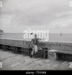 1940, historische, eine gentlleman Rauchen einer Pfeife auf eine Bank, die an den Blick auf den Ozean in Swanage, Isle of Purbeck, England sitzen. Stockfoto