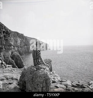 1940, historische, eine Dame sitzt auf einem Felsen über dem Meer in der Nähe der Tilly Laune Höhlen bei Swanage, auf der Isle of Purbeck, Dorset an der Jurassic Coast suchen. Diese alten Steinbrüchen abgebaut Purback Stein, eine Art von Kalkstein, der während der Napoleonischen Kriege für den Bau von Befestigungsanlagen verwendet wurde und Sie im Jahre 1887 als touristische Attraktion. Stockfoto