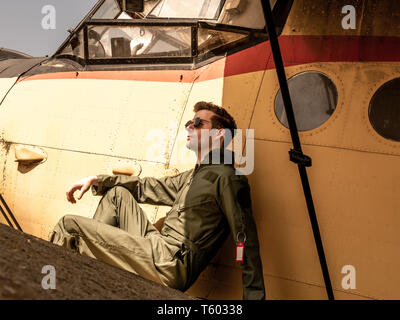 Ein hübscher junger Mann Pilot in einem grünen Insgesamt sitzen auf dem Flügel eines alten Platanen an einem sonnigen Tag. Stockfoto