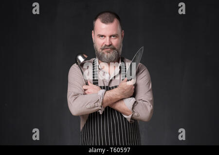 Reifen europäischen Chef mit Bart mit einer Schöpfkelle auf Schwarz studio Wand. Stockfoto