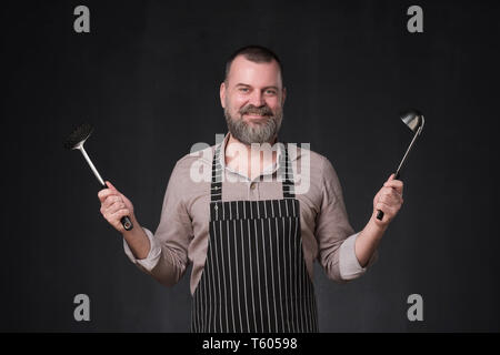 Reifen europäischen Chef mit Bart mit einer Schöpfkelle auf Schwarz studio Wand. Stockfoto