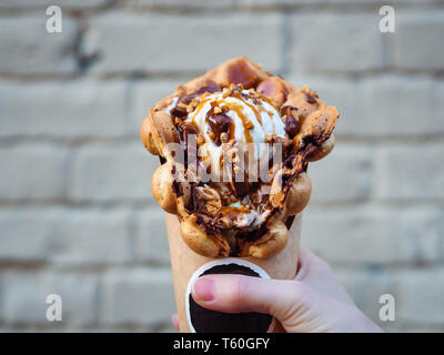 Hong Kong Waffeln in weiblicher Hand auf Mauer Hintergrund. Köstliche bubble Waffeln mit Eis und Karamell Sirup in der Frau die Hand. Trendy essen. Stockfoto