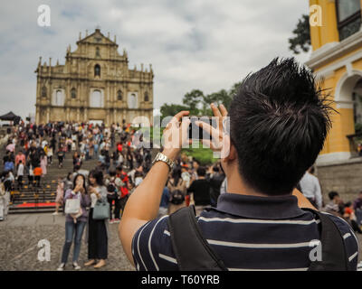 November 2018 - Macao, China: Junge asiatischer Mann mit einem Smartphone in eine grosse Masse der chinesischen Touristen ein Bild von der Ruinen von St. Paul's zu nehmen Stockfoto